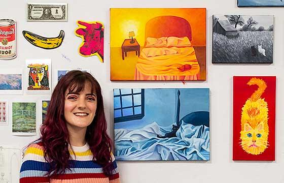 woman with long hair and bangs sitting in front of bright paintings hung on wall